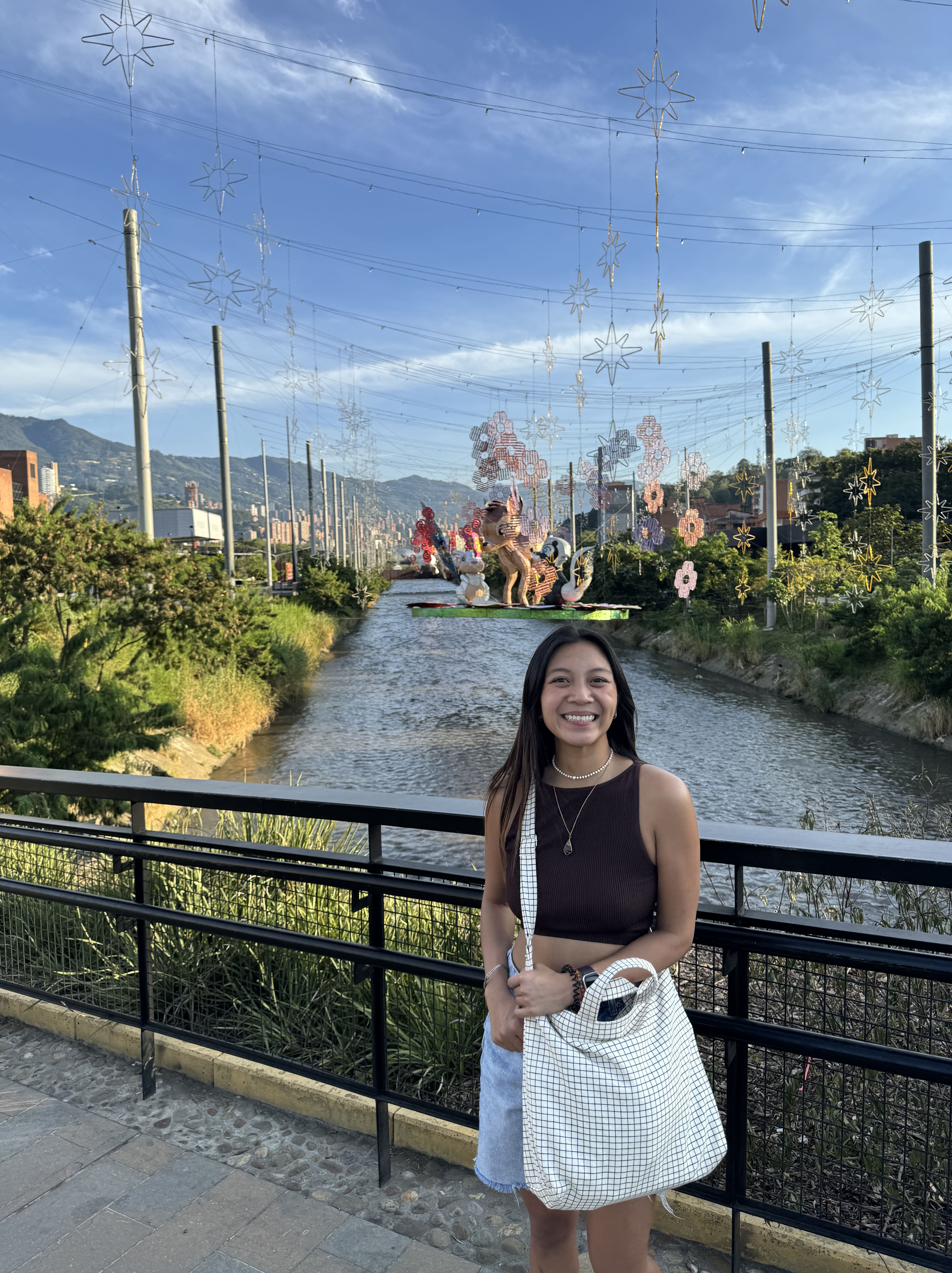 Girl posing in front of a river with lights and holding her bag on a nice day outside in Laureles at the park river walk 
